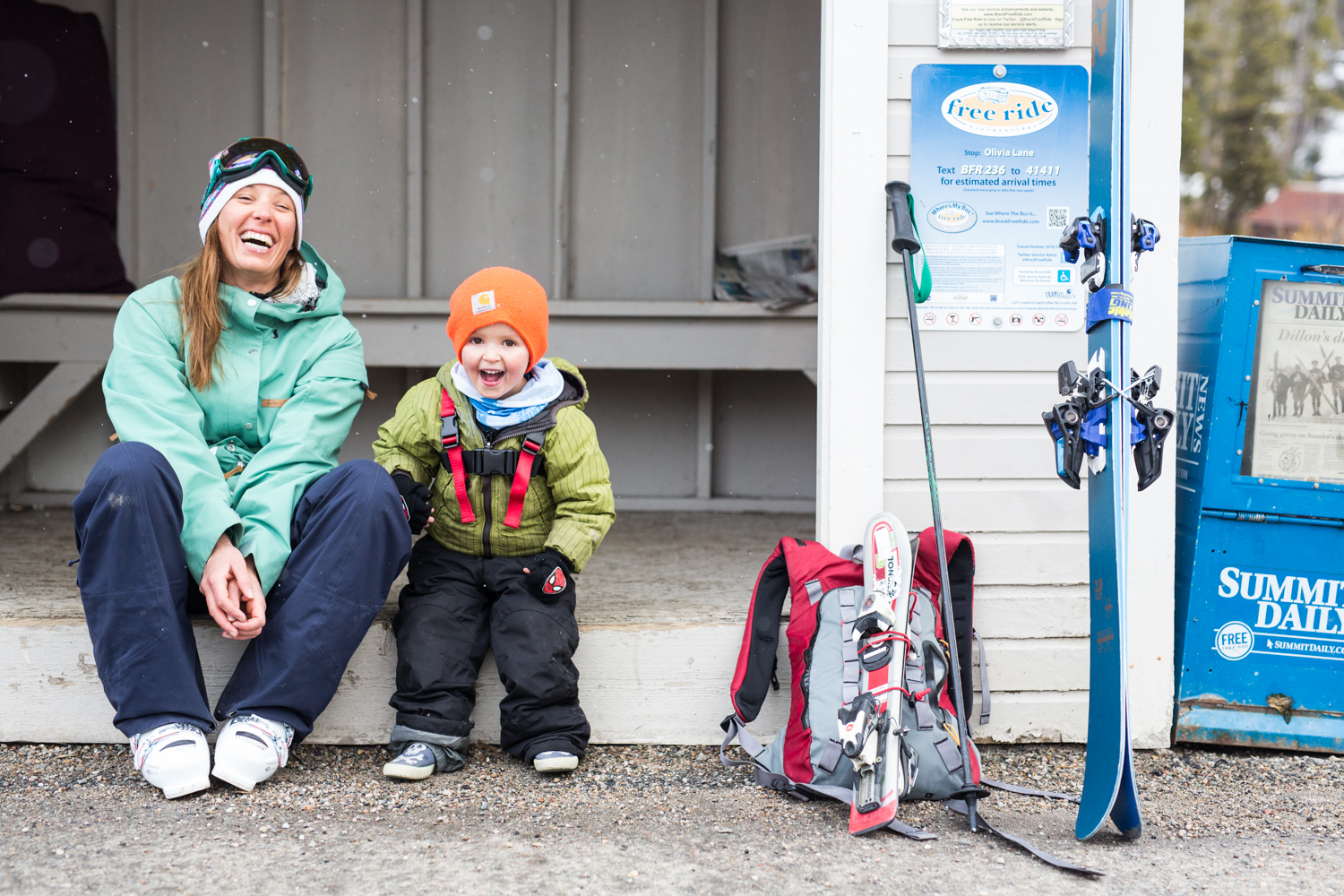 Parent and child smiling and sitting next to ski gear.